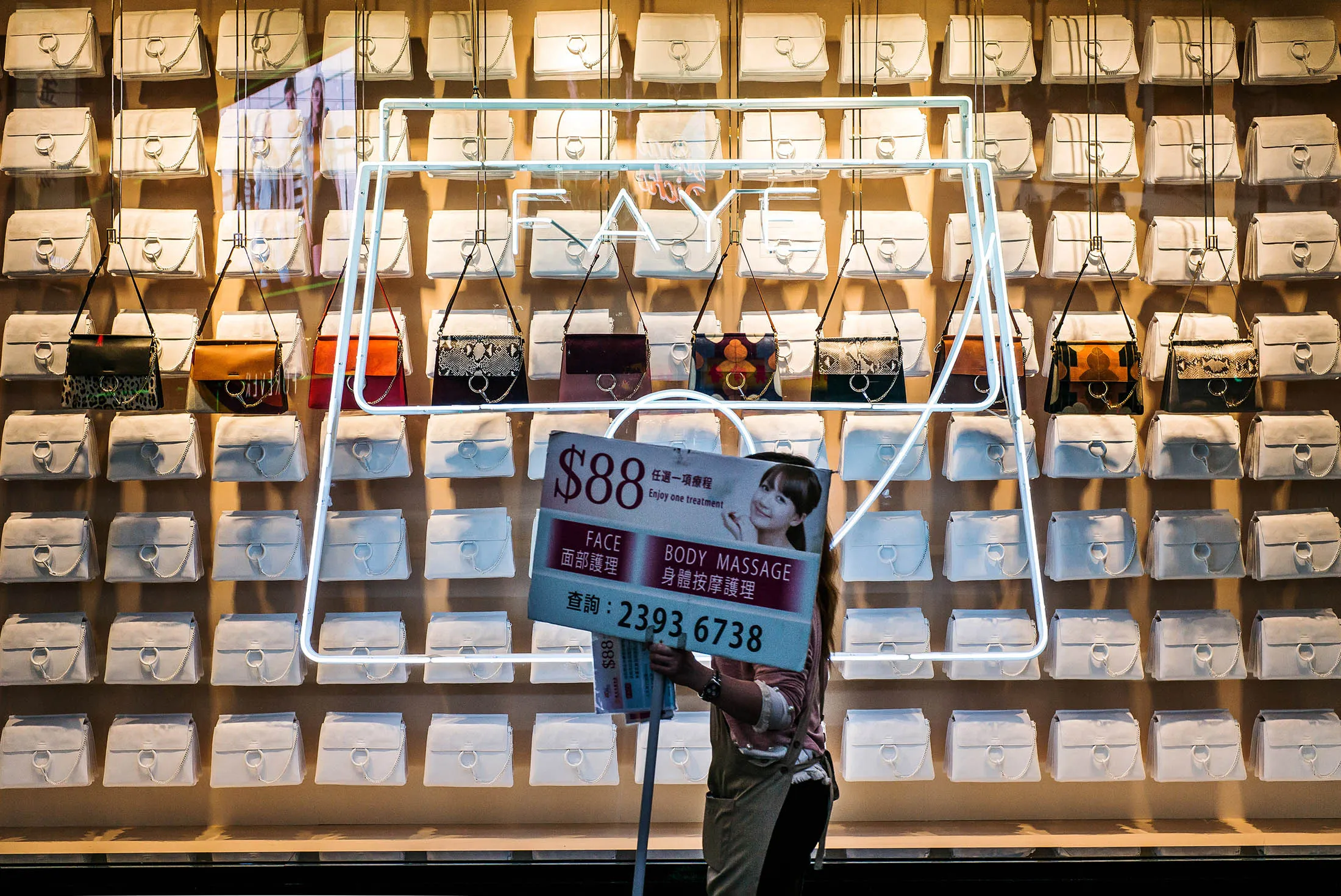 A human billboard carrying an advertising placard for body treatments walks past handbags in a window display of a store at night in the Causeway Bay district of Hong Kong, China, on Thursday, April 14, 2016. Hong Kong is scheduled to release consumer price index (CPI) figures on April 21.
