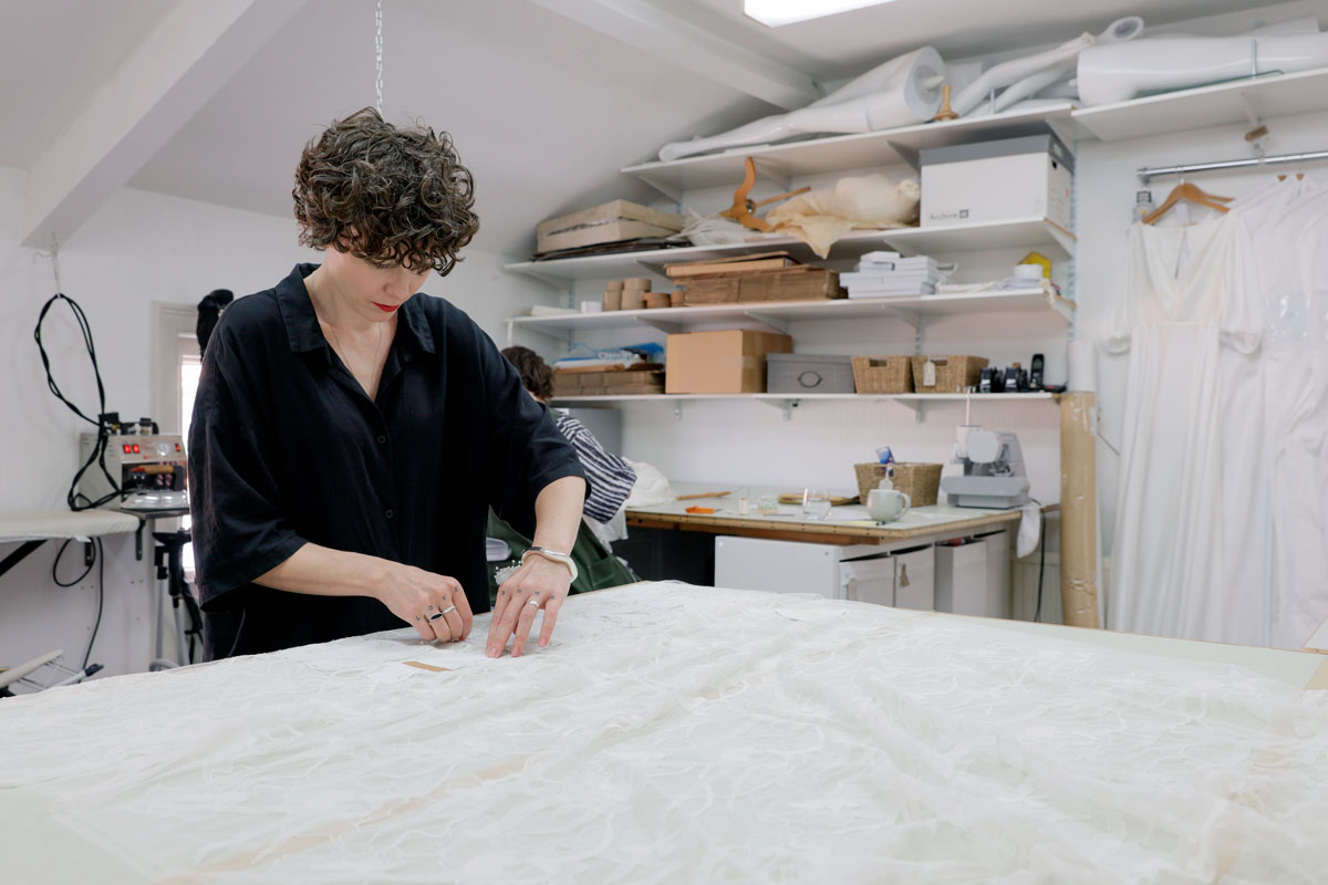A York-based wedding dressmaker is standing in front of a table and pinning down a pattern made of ivory lace fabric.