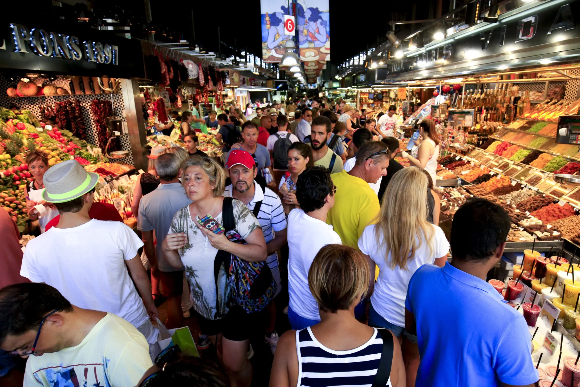 Tourists and pedestrians fill an aisle between food stalls inside Boqueria market in Barcelona, Spain.