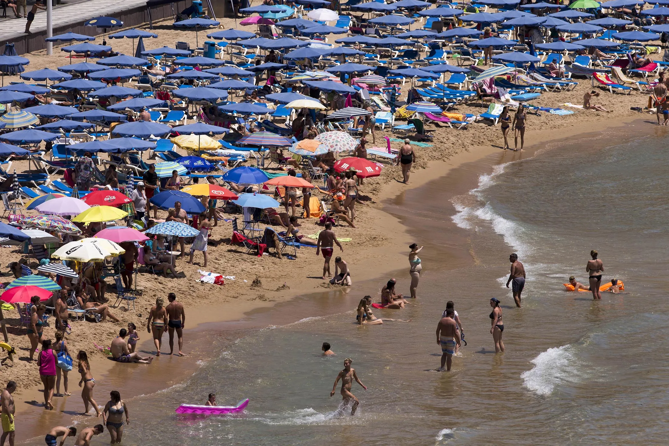 Holidaymakers sit beneath parasols and play in the surf on the beachfront in Benidorm, Spain, on Tuesday, July 12, 2016. Spain registered record tourism year in 2015 with 68 million visitors.
