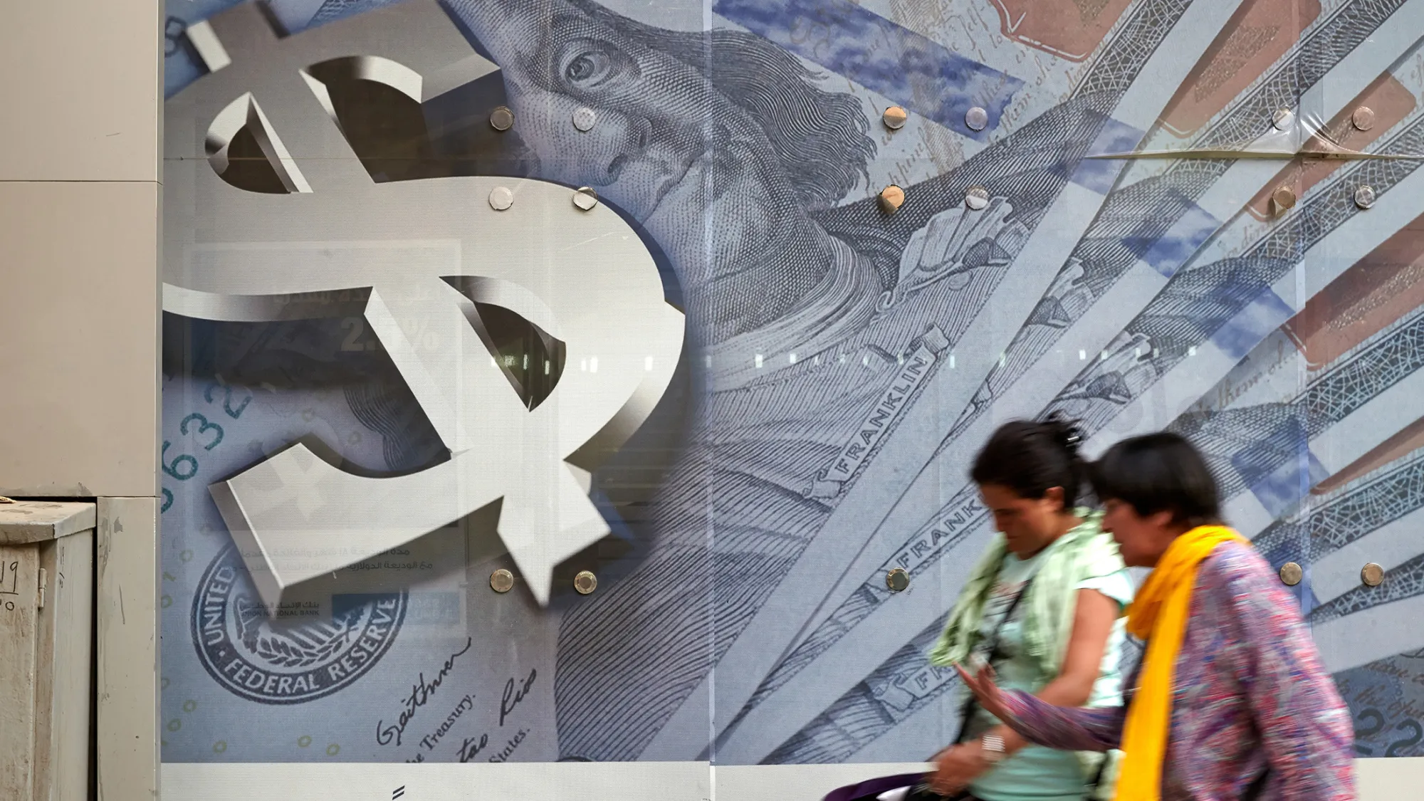 Pedestrians walk past an advertisement displaying a large dollar sign and U.S. dollar banknotes.
