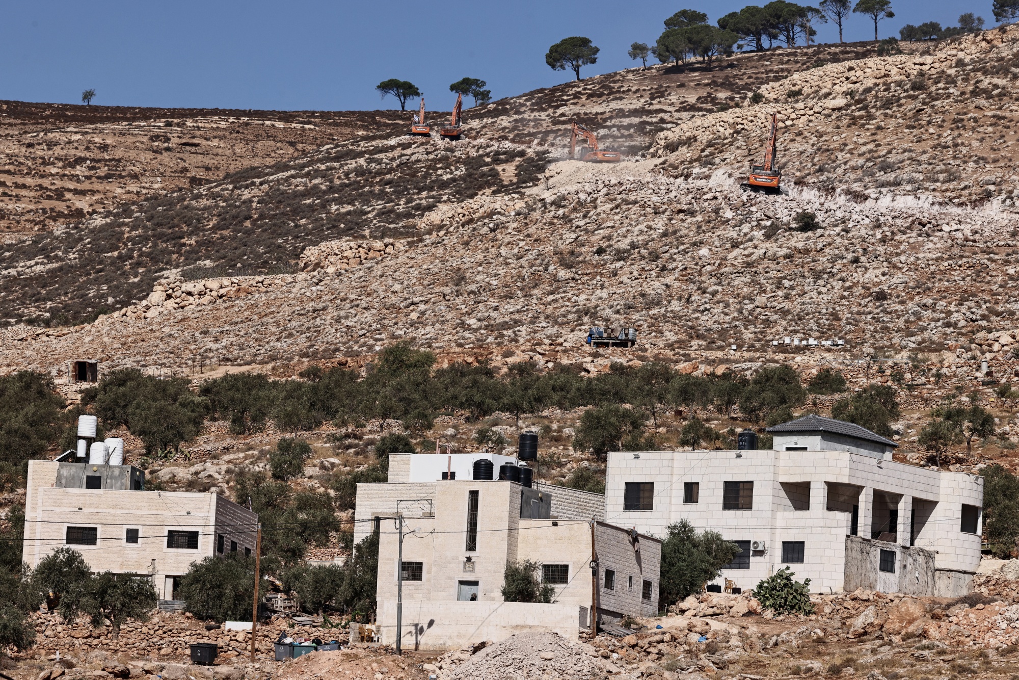 Several bulldozers work on a rocky hillside to carve a road for a new settlement near Al-Mughayyir, north of Ramallah. Modern stone houses with water tanks on their rooftops sit in the foreground.