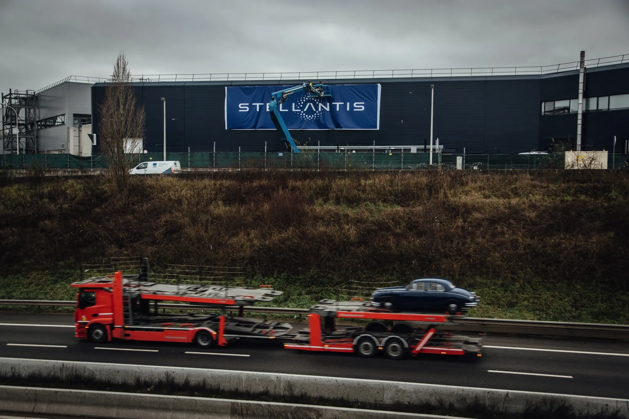 A vehicle transporter passes workers installing a Stellantis NV logo banner&nbsp;in Velizy-Villacoublay,&nbsp;France.