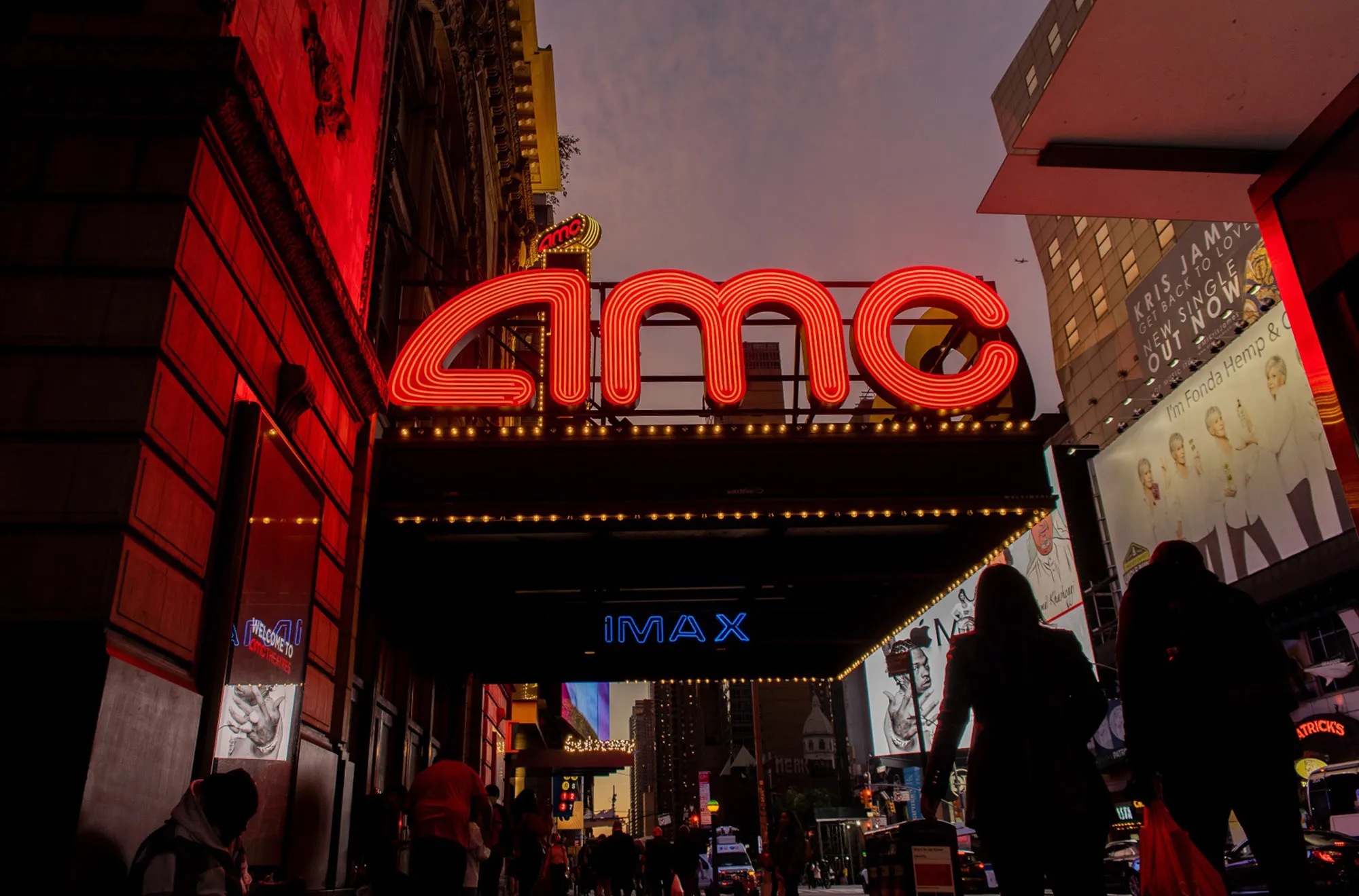 Pedestrians pass in front of an AMC movie theater at night in the Times Square neighborhood of New York.