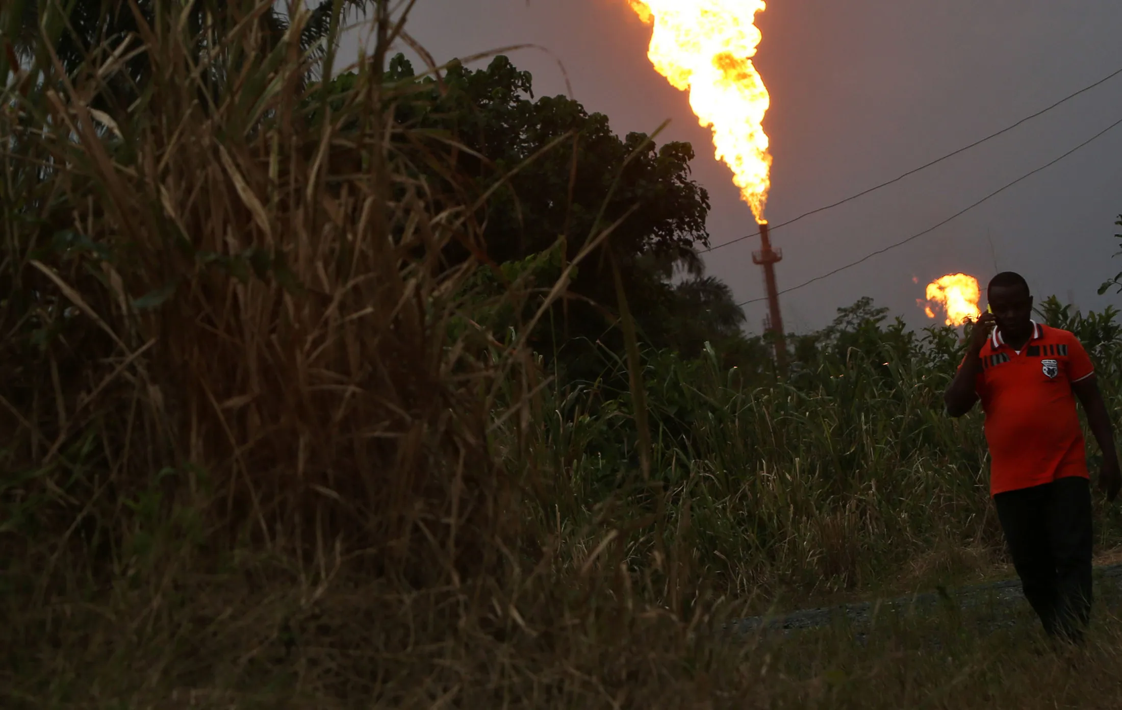 A man walks through foliage as gas flares burn in the distance at an oil flow station&nbsp;in Idu, Rivers State, Nigeria.