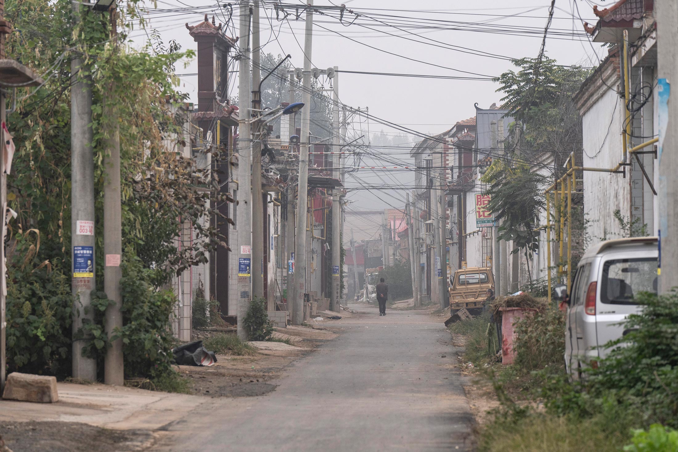 A man in a black suit walks through an alleyway with many electrical wires. The scene is smoggy. 