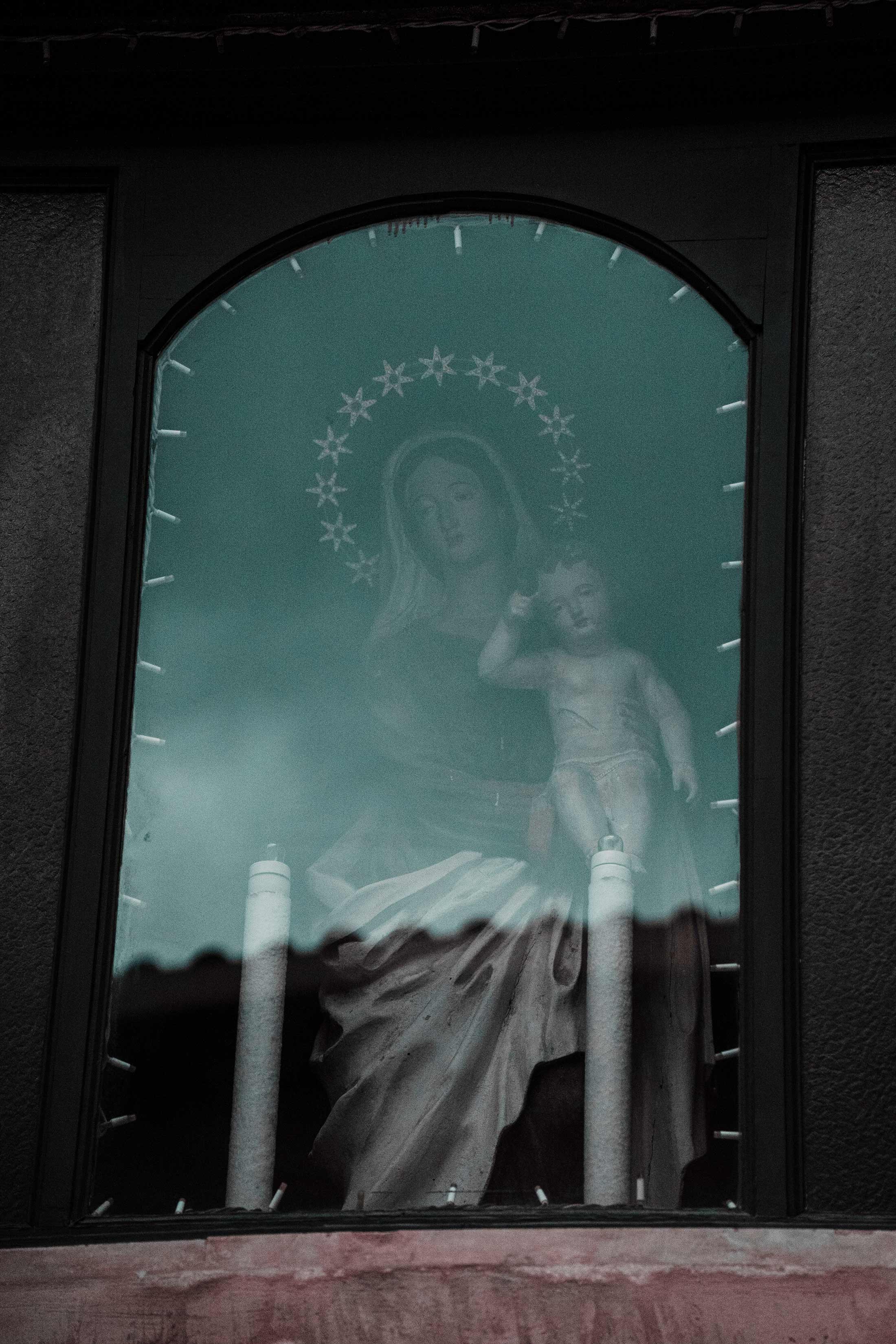 A statue of the virgin Mary with stars around her head as seen through a church window. She is holding the baby Jesus. 
