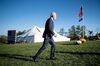 Biden, wearing a suit and tie and a face mask, walks alone on a grassy field. Behind him is a large covered white tent and two large American flags, one on a board in front of the tent and the other on a flagpole.