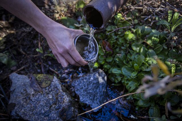 An environmentalist fills a cup with spring water from a pipe beside one of the stone structures where the company BlueTriton Brands collects water and sends it flowing through pipes across the San Bernardino National Forest.