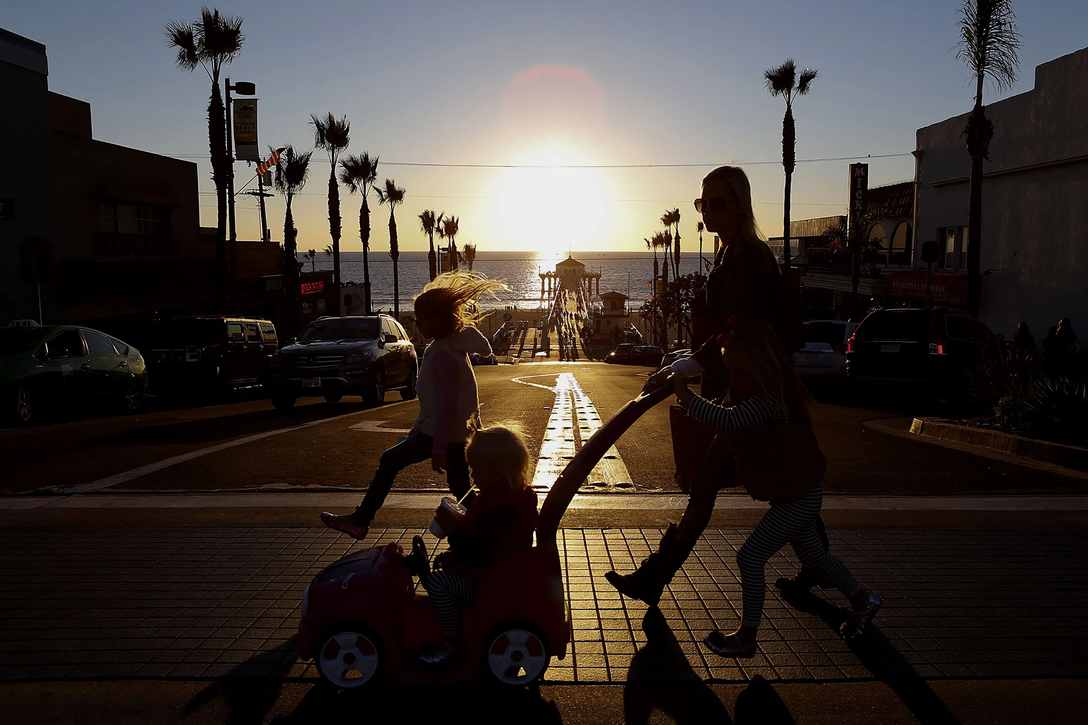 A woman and three children cross the street at sunset near storefronts by the pier in Manhattan Beach, California.
