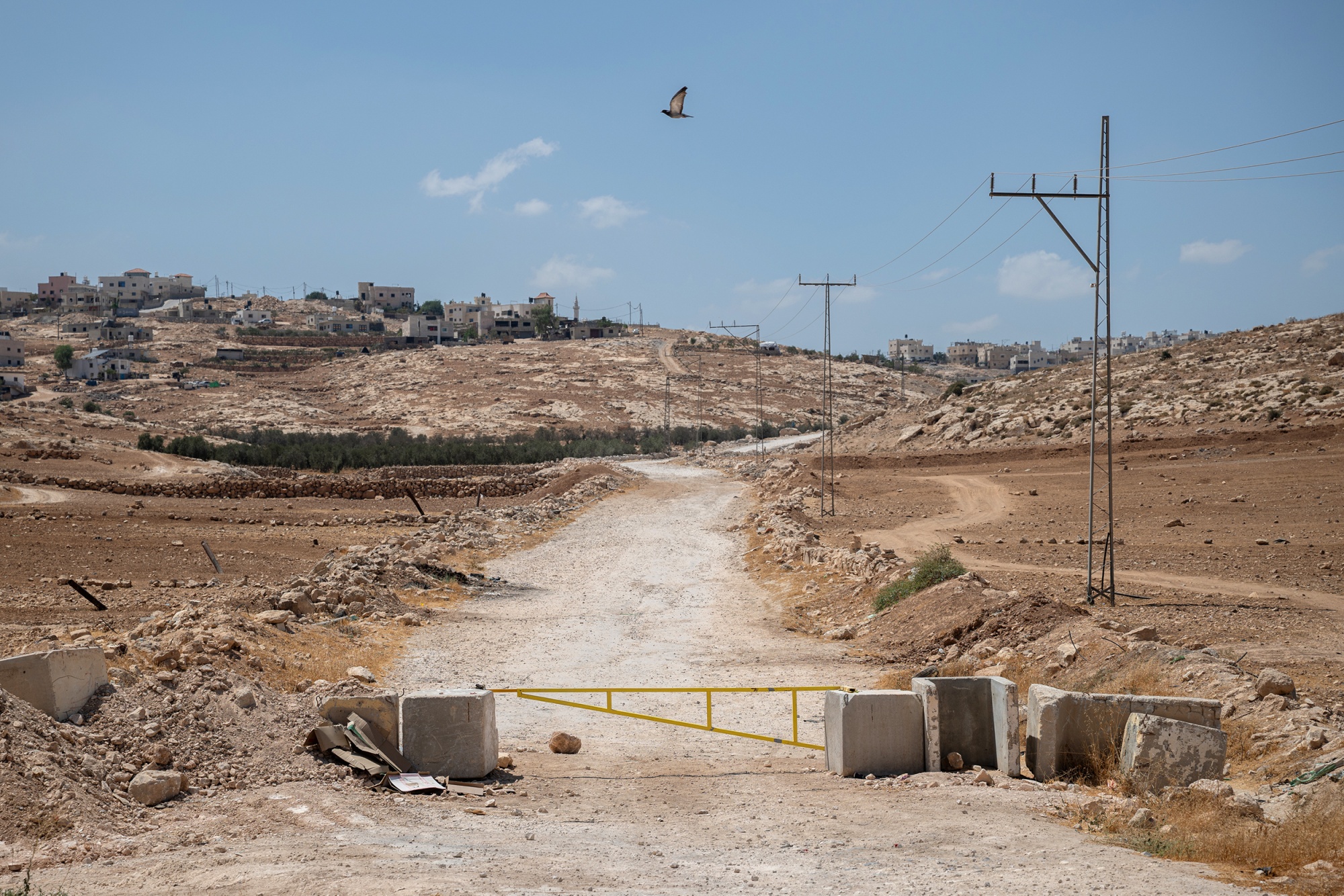 A yellow metal gate, reinforced by concrete slabs, blocks a dirt road leading toward the Palestinian village of Al-Jwaya in the South Hebron Hills, restricting residents' access to the main highway, with scattered homes visible on the hillside in the background.