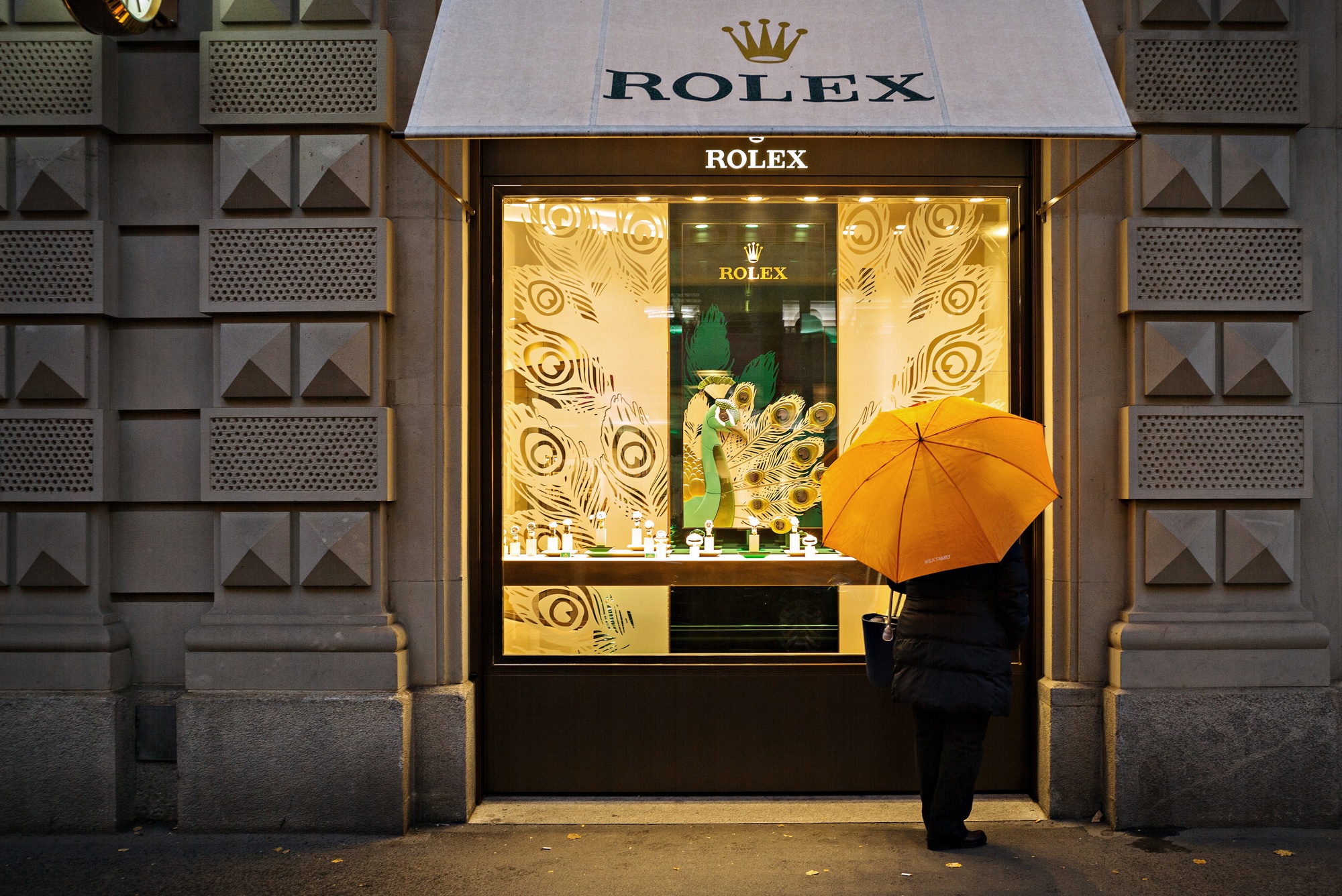 A pedestrian shelters under an umbrella whilst browsing the window of a Rolex Group luxury wristwatch store in Zurich, Switzerland
