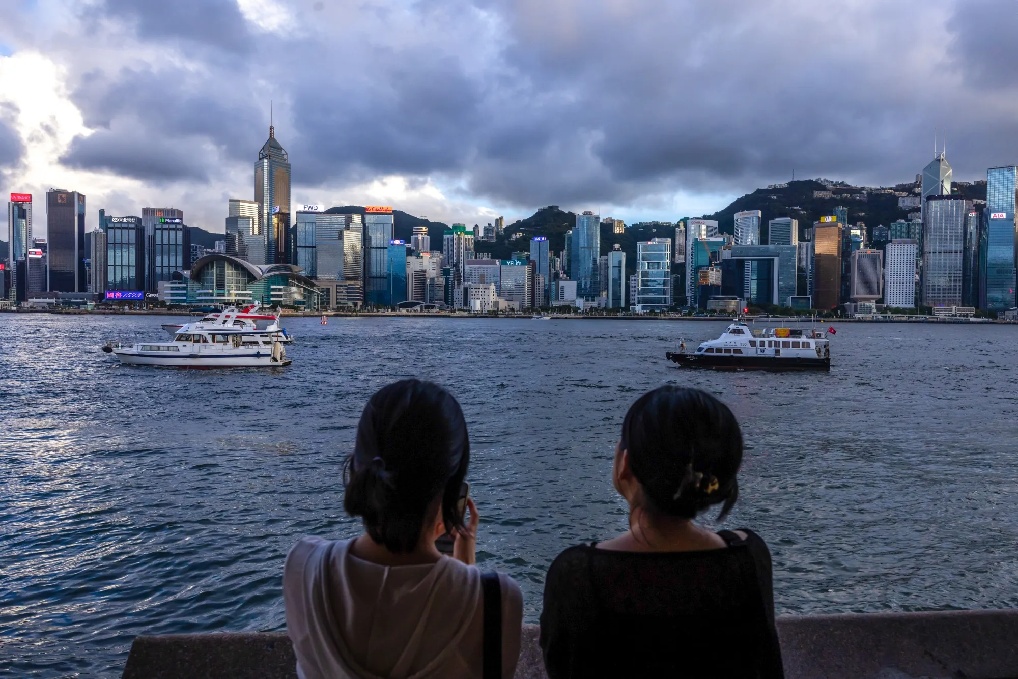 Pedestrians look out across Victoria Harbour towards the Hong Kong Island skyline in the Tsim Sha Tsui district of Hong Kong, China.