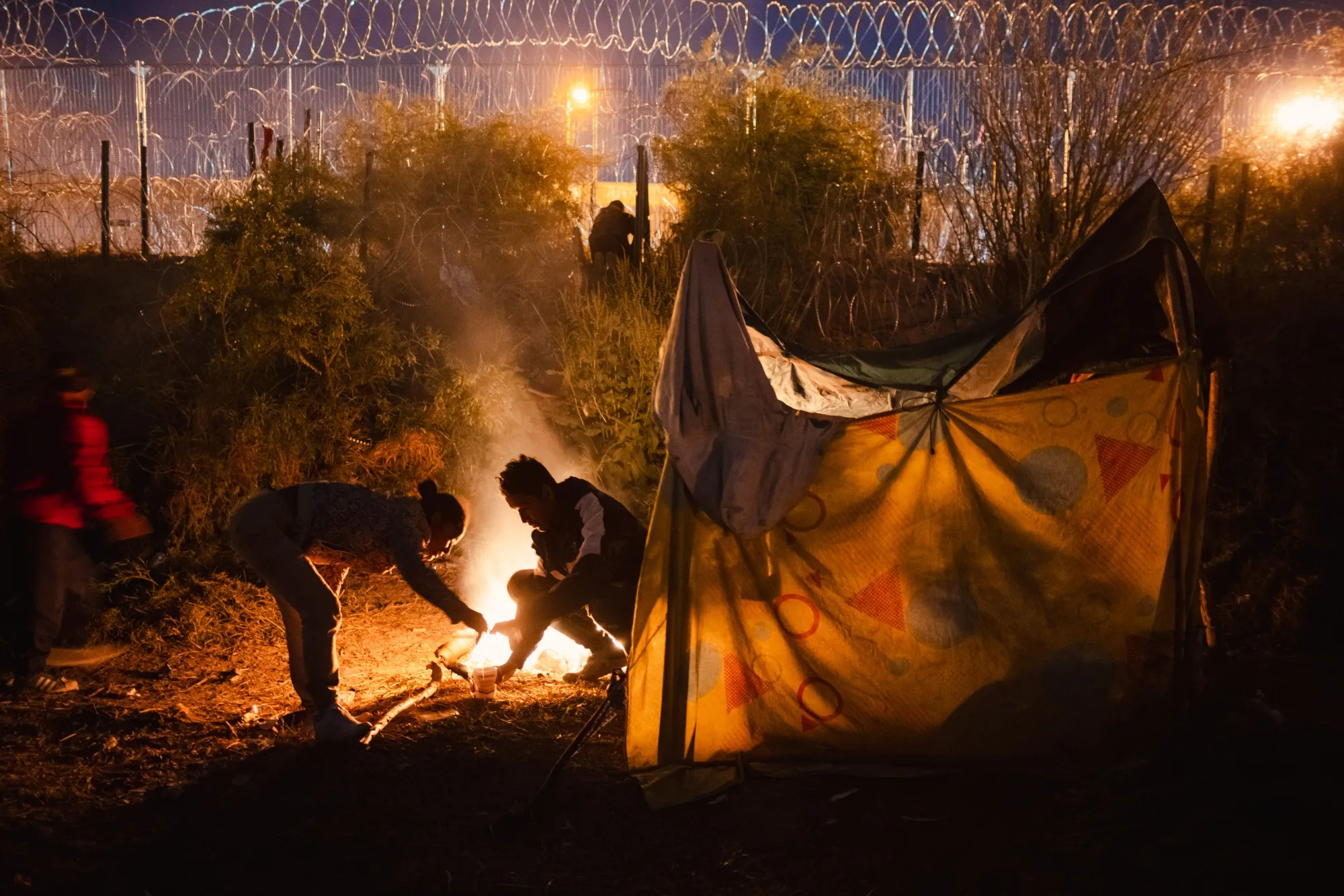 A migrant family cooks dinner over a fire as another migrant, top center, cuts through concertina wire after crossing the border in El Paso, Texas, on April 3.