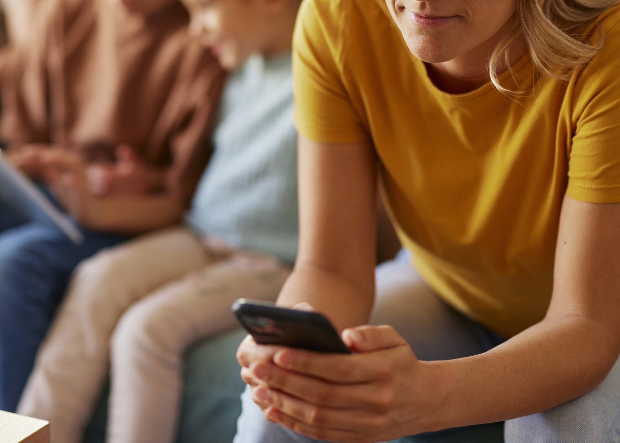 Carefree mother text messaging on smart phone while relaxing next to her family on sofa in the living room. RF