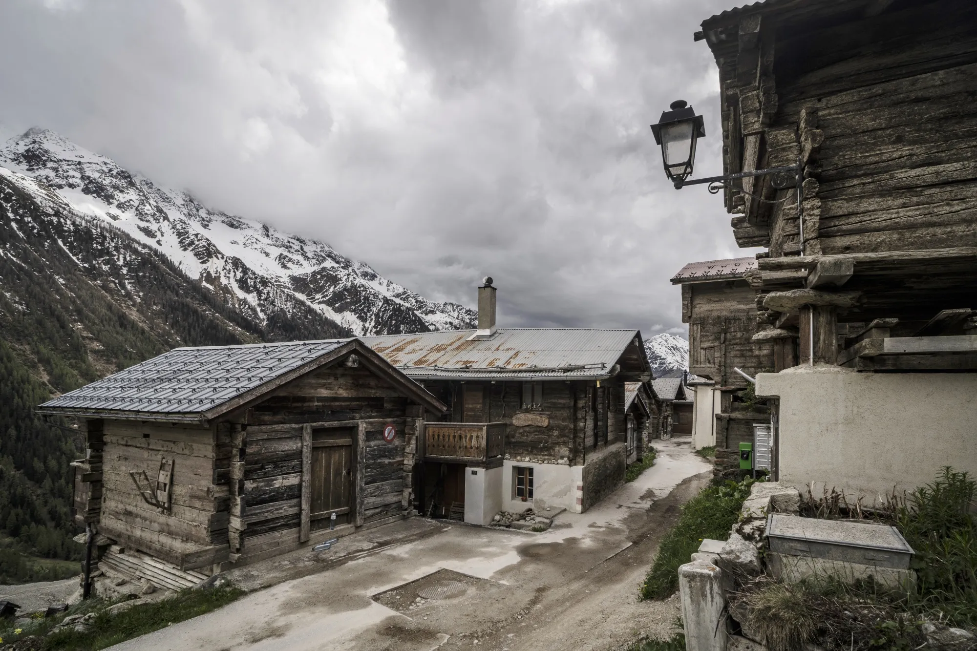 Wooden Swiss homes line an empty street. In the background dark storm clouds fill the valley and one steep moutain peak, capped by snow, is visible in the distance.