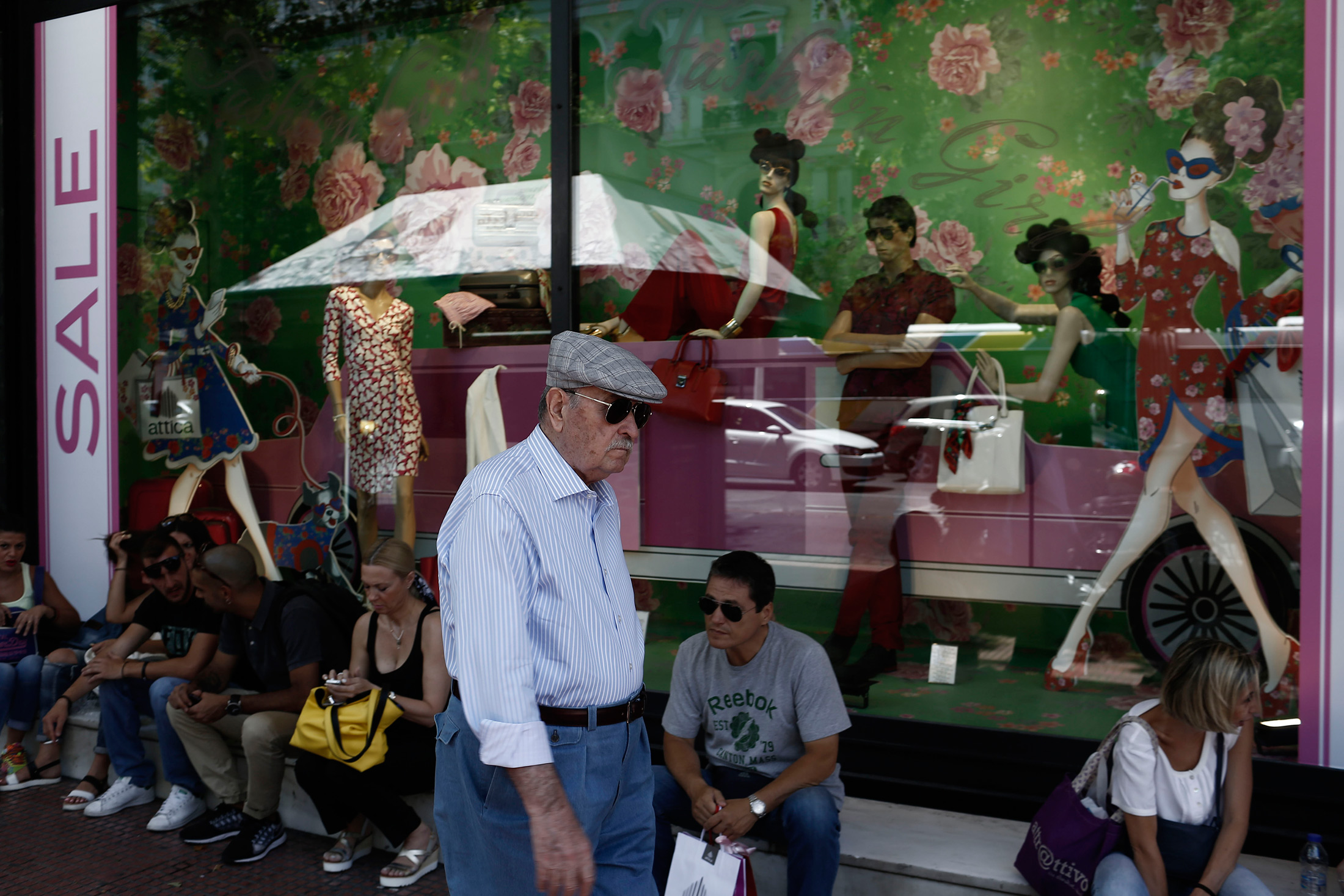 An older man passes pedestrians sitting outside a fashion store advertising price discounts in the summer sales in Athens, Greece, on July 13, 2015.
