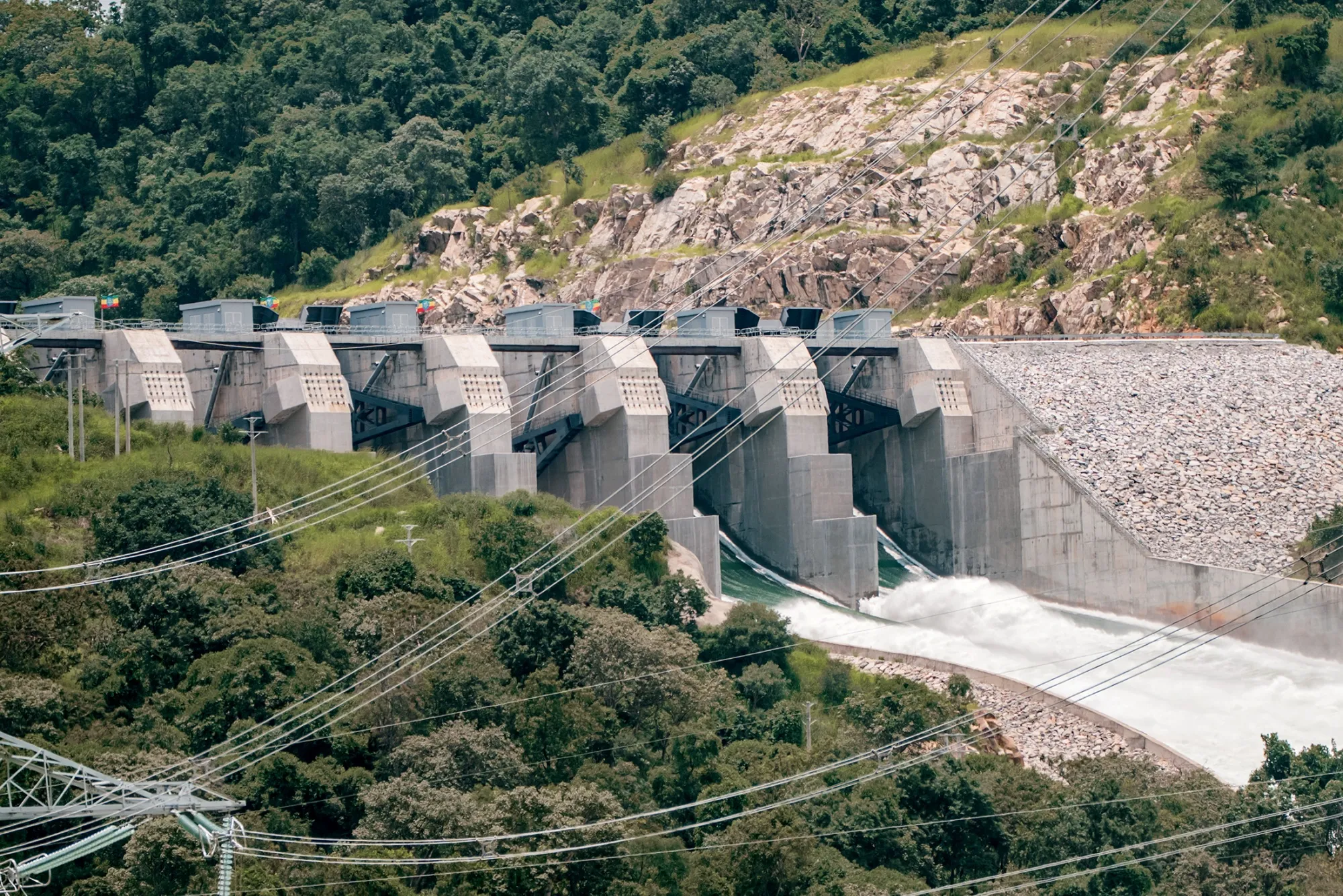 The Grand Ethiopian Renaissance Dam in Guba, Ethiopia, releases water through its spillways, surrounded by green hills.