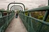 Person with cane and a shopping barrow walking on a steel bridge.