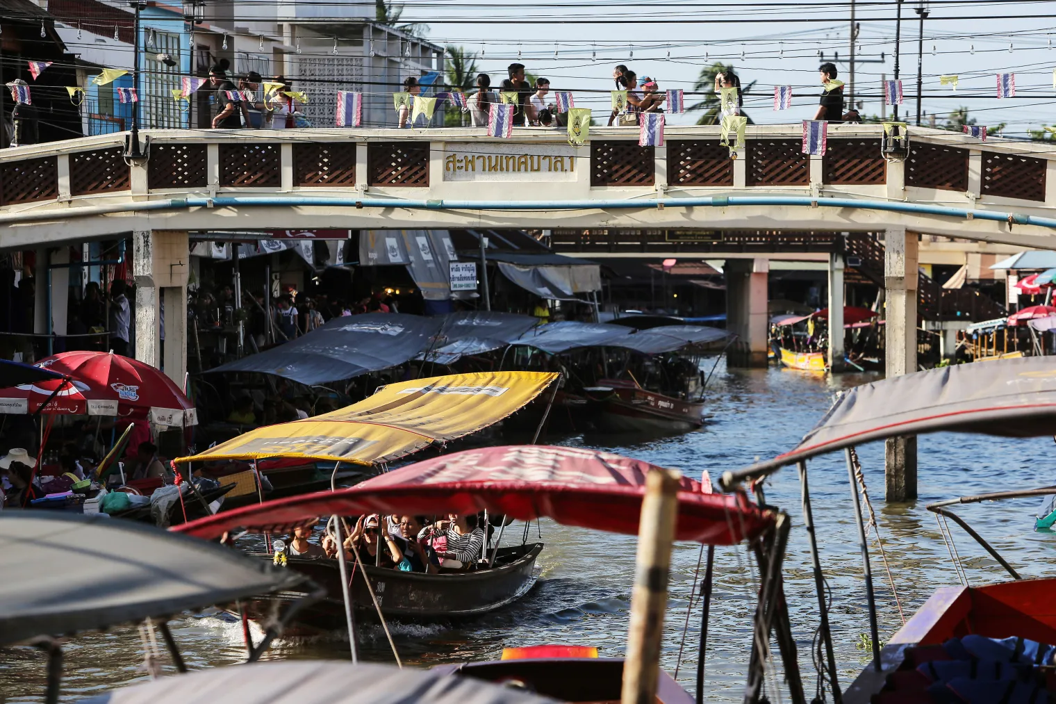 Pedestrians walk along a bridge as passengers ride on a boat at the Amphawa Floating Market in Aphawa, Samut Songkhram Province, Thailand.