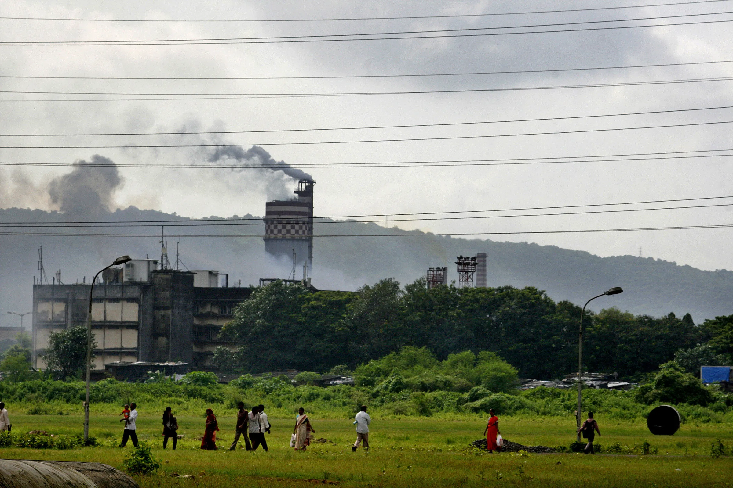 A people walk down a road in front of a chemical factory as it discharges exhaust into the air of Mumbai, India