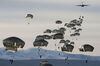 US Army airborne troops parachute from a plane in the distance, like a bloom of jellyfish being littered from the sky.