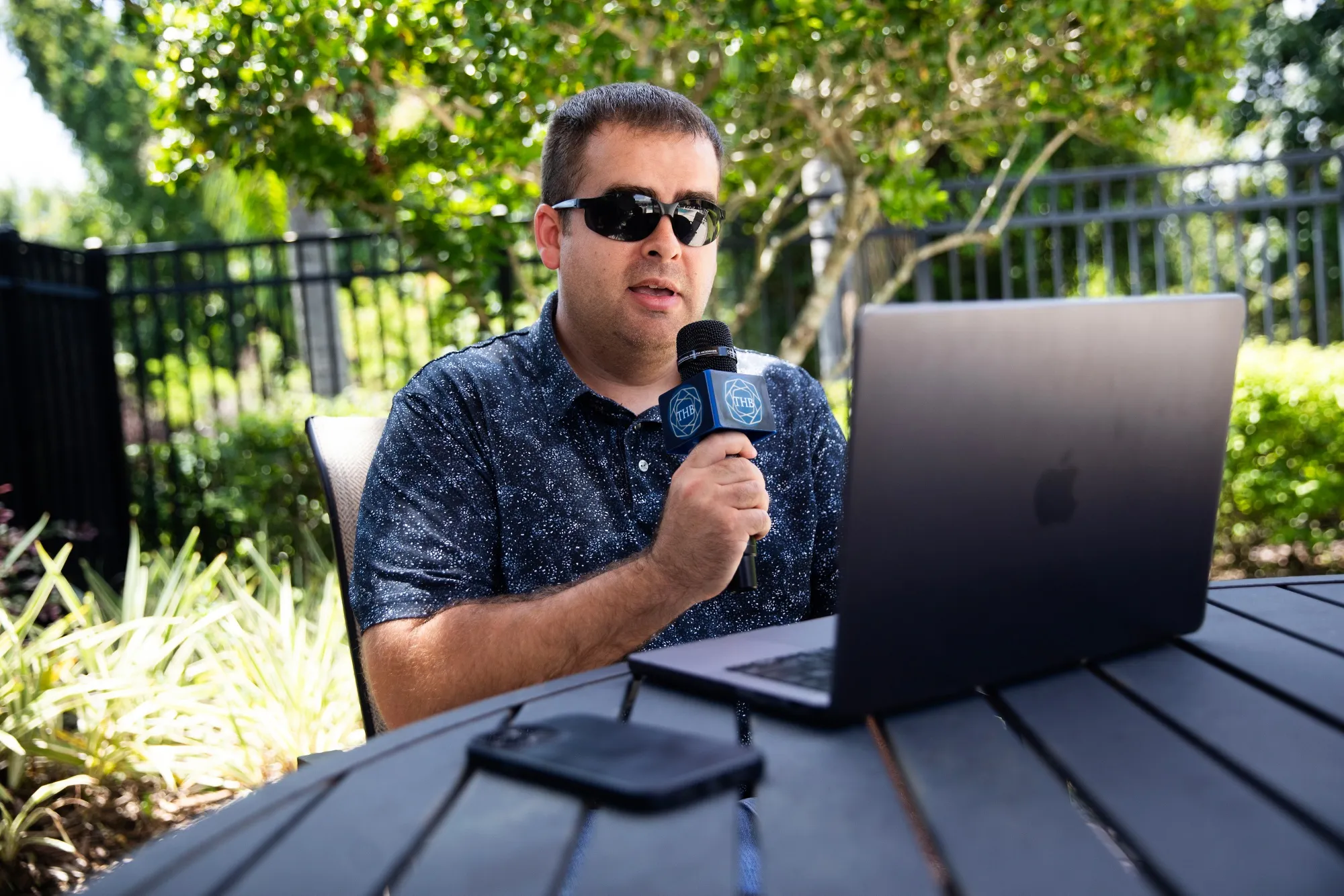 Thomas Bell speaks into a microphone with his initials "THB" while sitting outside in front of a laptop.