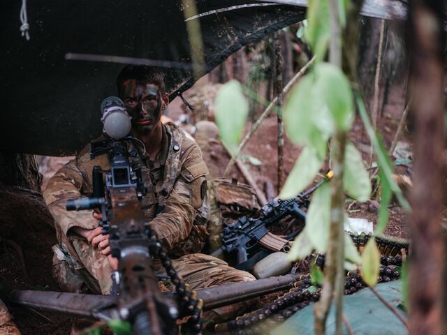 A solidier in army fatigues and camouflage face paint surrounded by weapons looks at the camera.