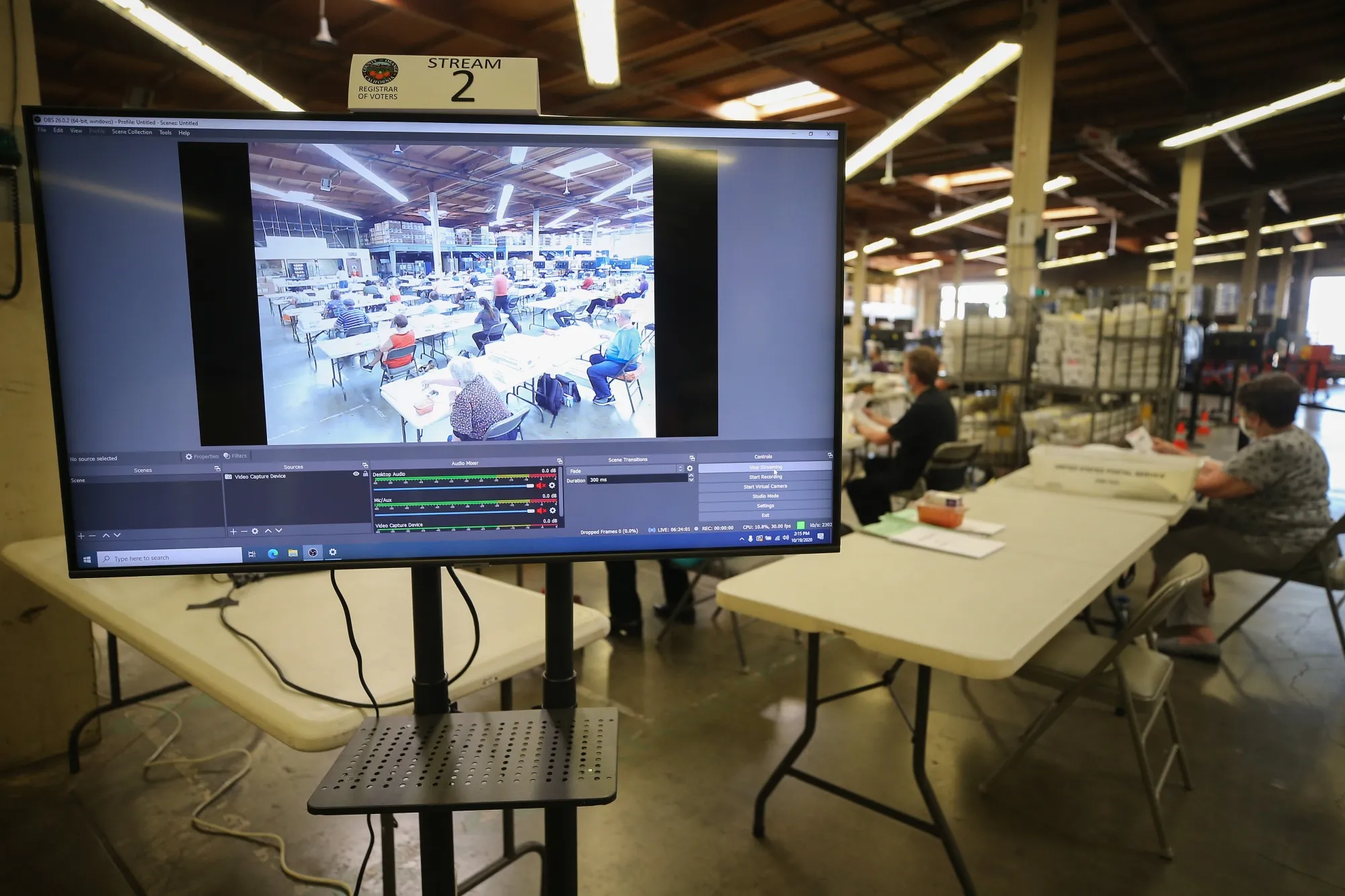 Election workers process mail-in ballots as a monitor displays a livestream of the process at the Orange County Registrar of Voters on Oct. 19, 2020 in Santa Ana, California. It’s one of a number of counties livestreaming their ballot counts this year to bolster faith in the integrity of the process.&nbsp;