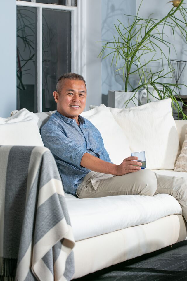 Simpson wong seated on white sofa at home. Home contains plant in background and Simpson is holding a drinking glass.