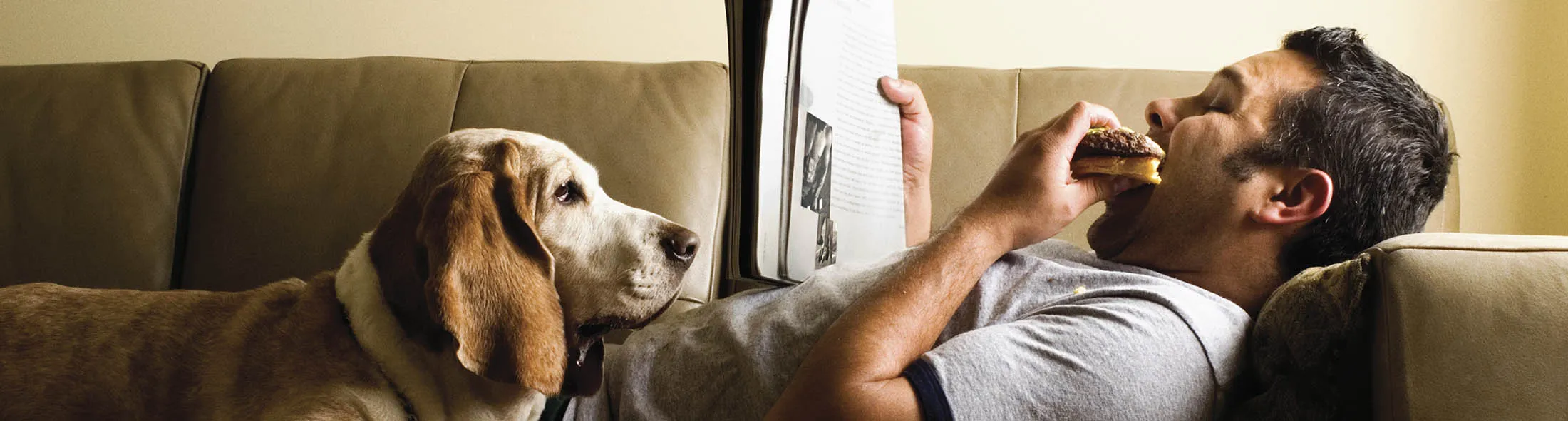 Man lying on sofa with dog, eating hamburger, reading magazine
