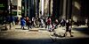 Pedestrians walking in a tunnel of sunlight along Broadway at the intersection with Wall Street in the financial district of New York City.