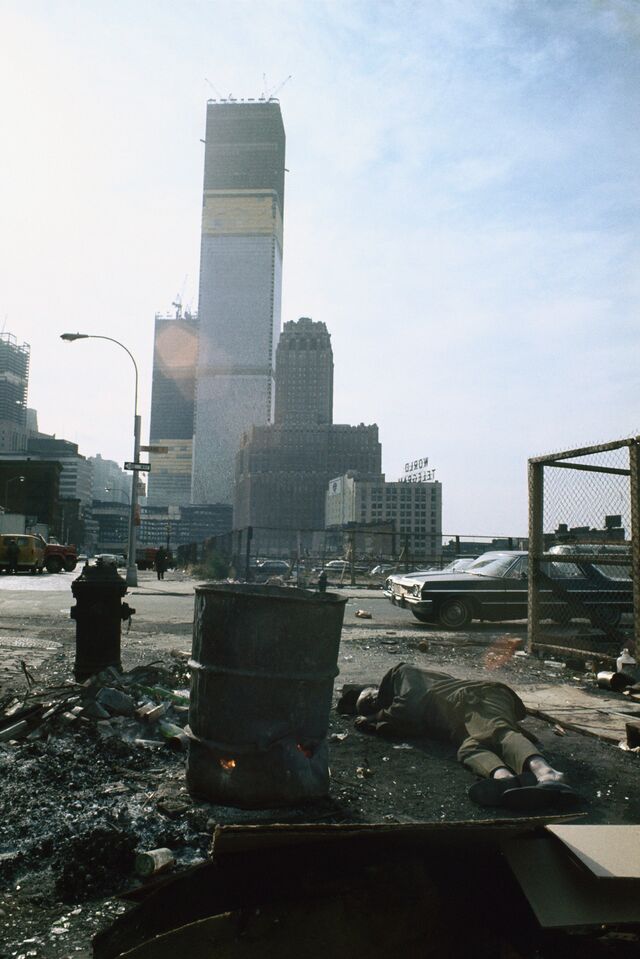 A view of the twin towers from Duane Street with person sleeping on the ground next to trash can, 1970.