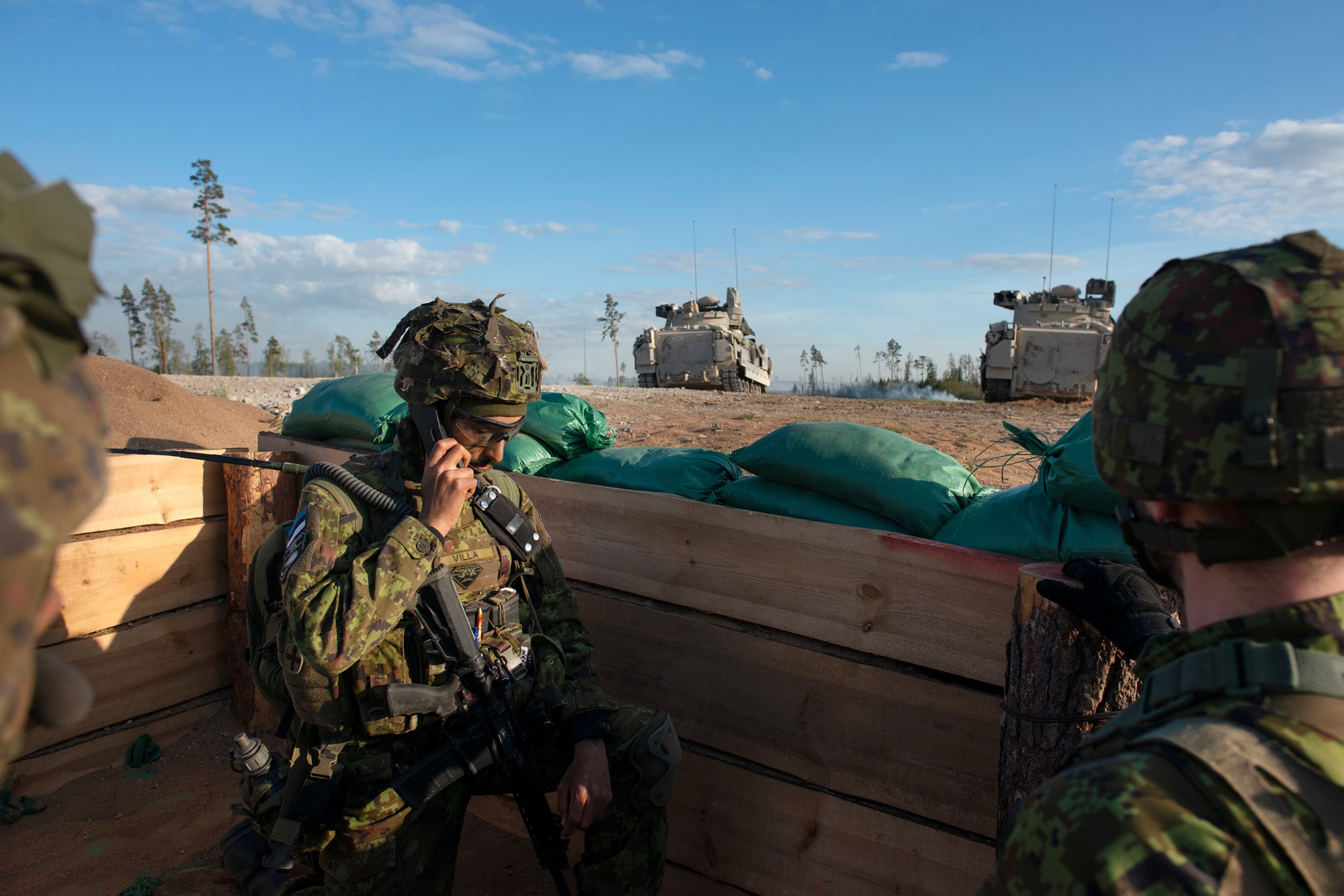 Two men in combat fatigues listen to a radio inside an open shelter. There are two tanks in the background.