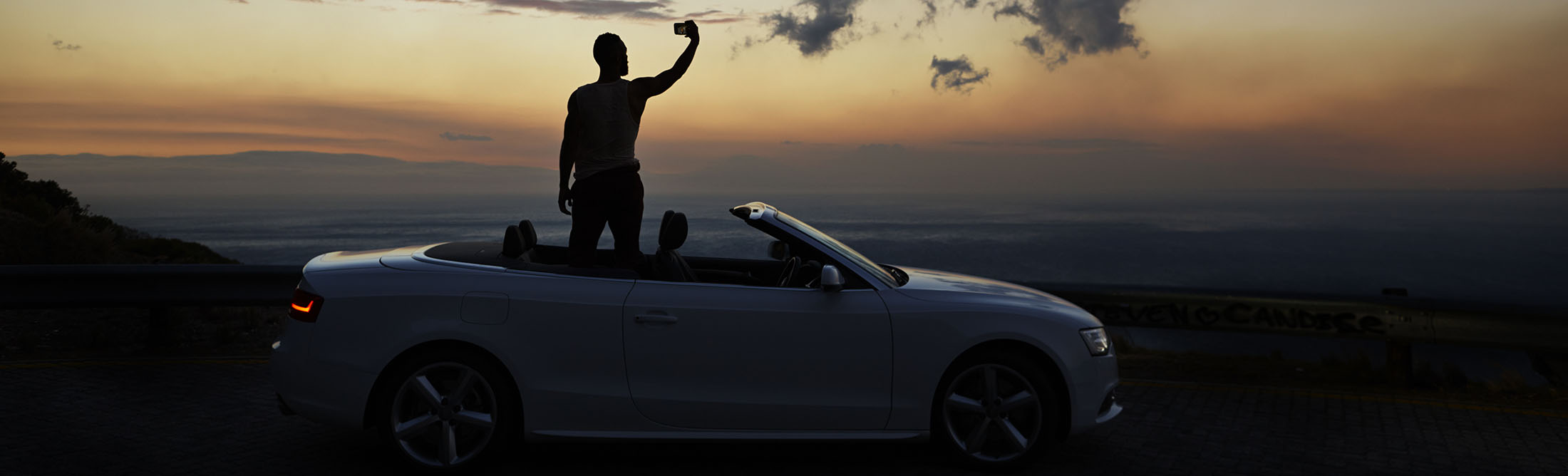 Man taking photo of sunset with smartphone, standing in convertible car