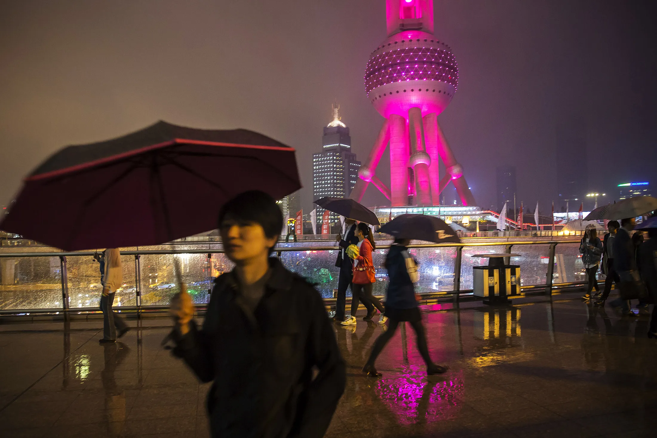 Pedestrians holding umbrellas walk as the Oriental Pearl Tower, right, stands illuminated at night in the Lujiazui district of Shanghai, China.
