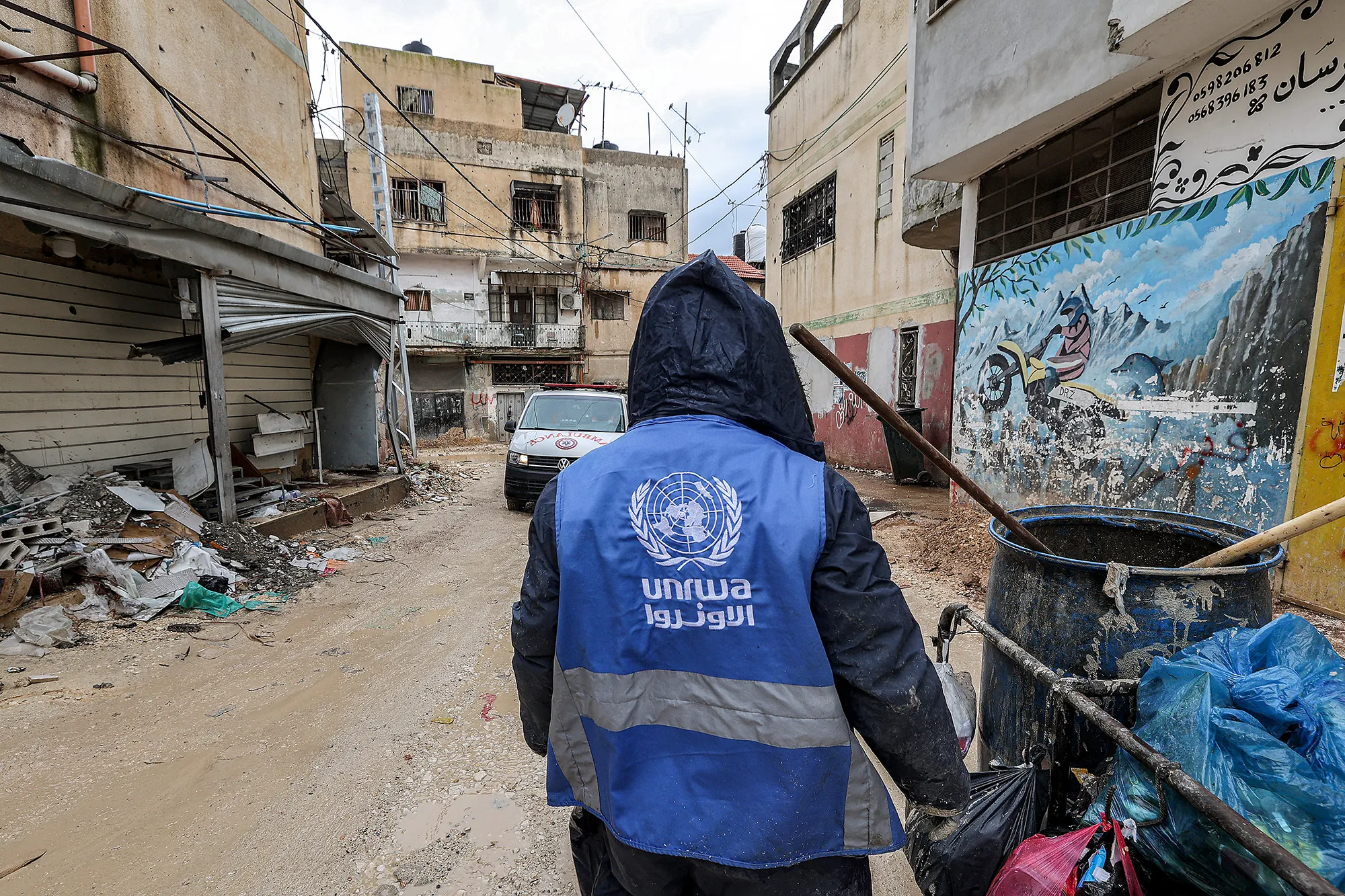 A man collects trash while wearing a jacket bearing the logo of the United Nations Relief and Works Agency for Palestine Refugees in Jenin, occupied West Bank.