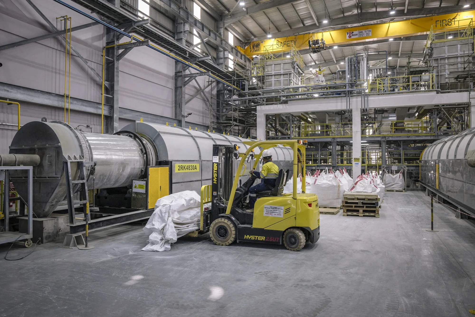 A worker drives a yellow forklift past large rotary kilns inside the Lynas Advanced Materials Plant.