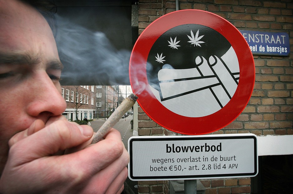 A man smokes a marijuana cigarette in front of a sign prohibiting marijuana smoking in public.