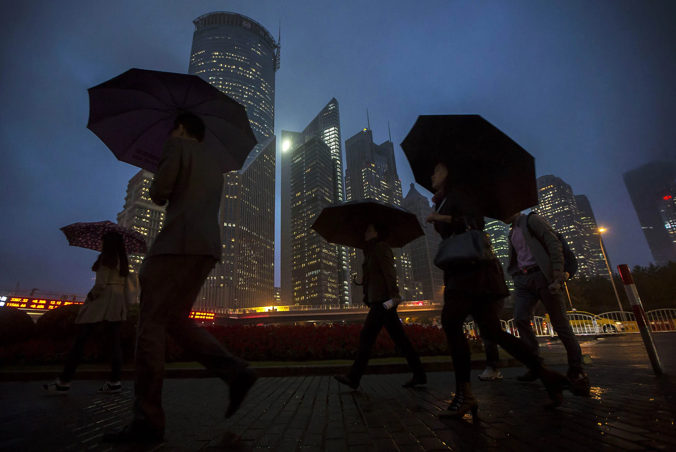 Pedestrians holding umbrellas walk past buildings illuminated at night in the Lujiazui district of Shanghai, China, on Thursday, Oct. 29, 2015. The Communist Party ended its four-day plenum on Thursday, with Premier Li Keqiang signaling the leadership may be ready to accept the weakest period of expansion since the economy was opened up three decades ago.
