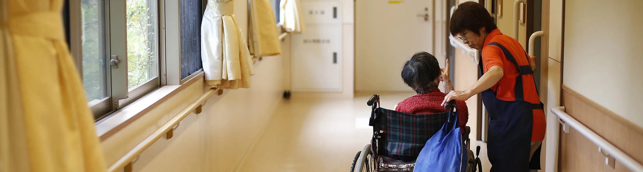 Sonoe Kudo, 65 year old caregiver, leads a resident riding on a wheelchair to her room at a nursing home operated by CARE TWENTY-ONE Corp. in Kasai, Tokyo, Japan, on Thursday, June 30, 2016. Bloomberg/Yuya Shino
