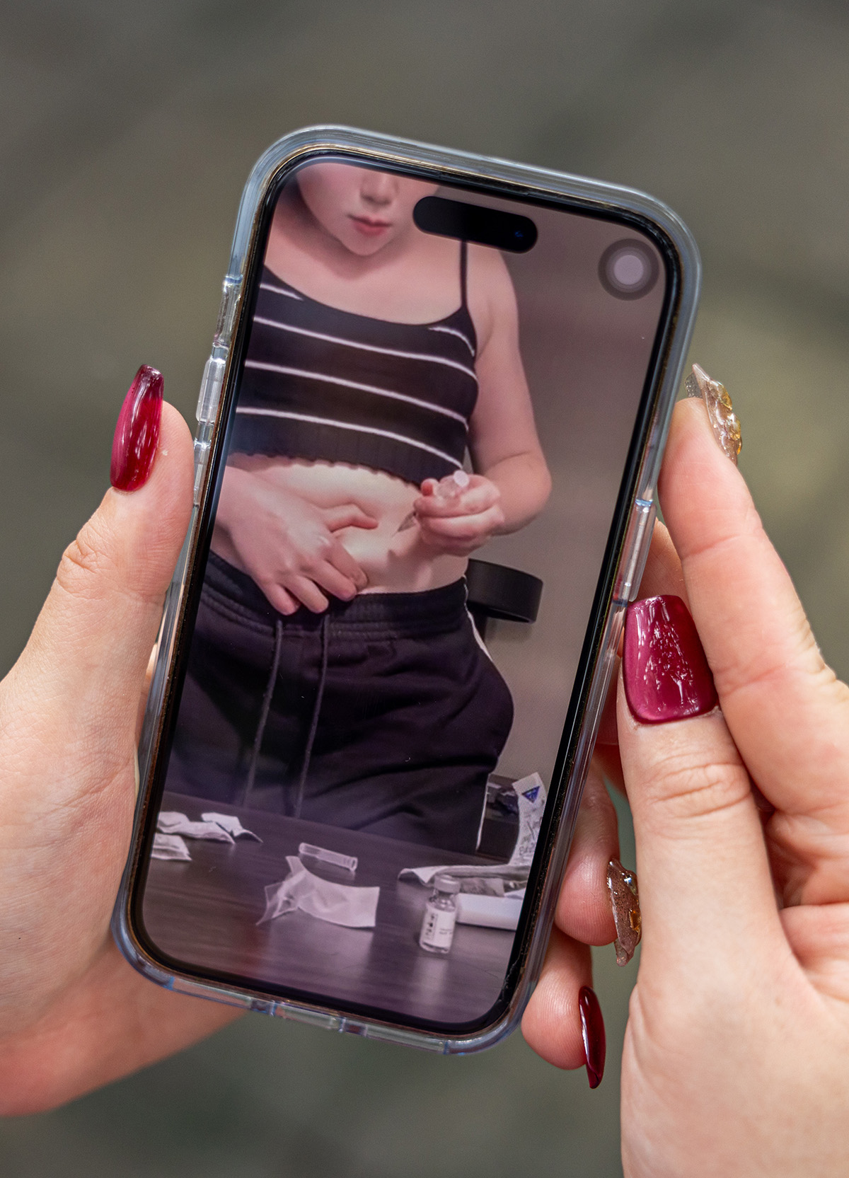 A woman with long pink nails holds a phone which shows another image of her wearing a cropped top and jeans while she injects herself in the abdomen with a needle. 