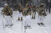 Five soldiers carrying heavy gear, two wearing sunglasses, pause with their ski poles in the ground. There are trees behind them and it is snowing.