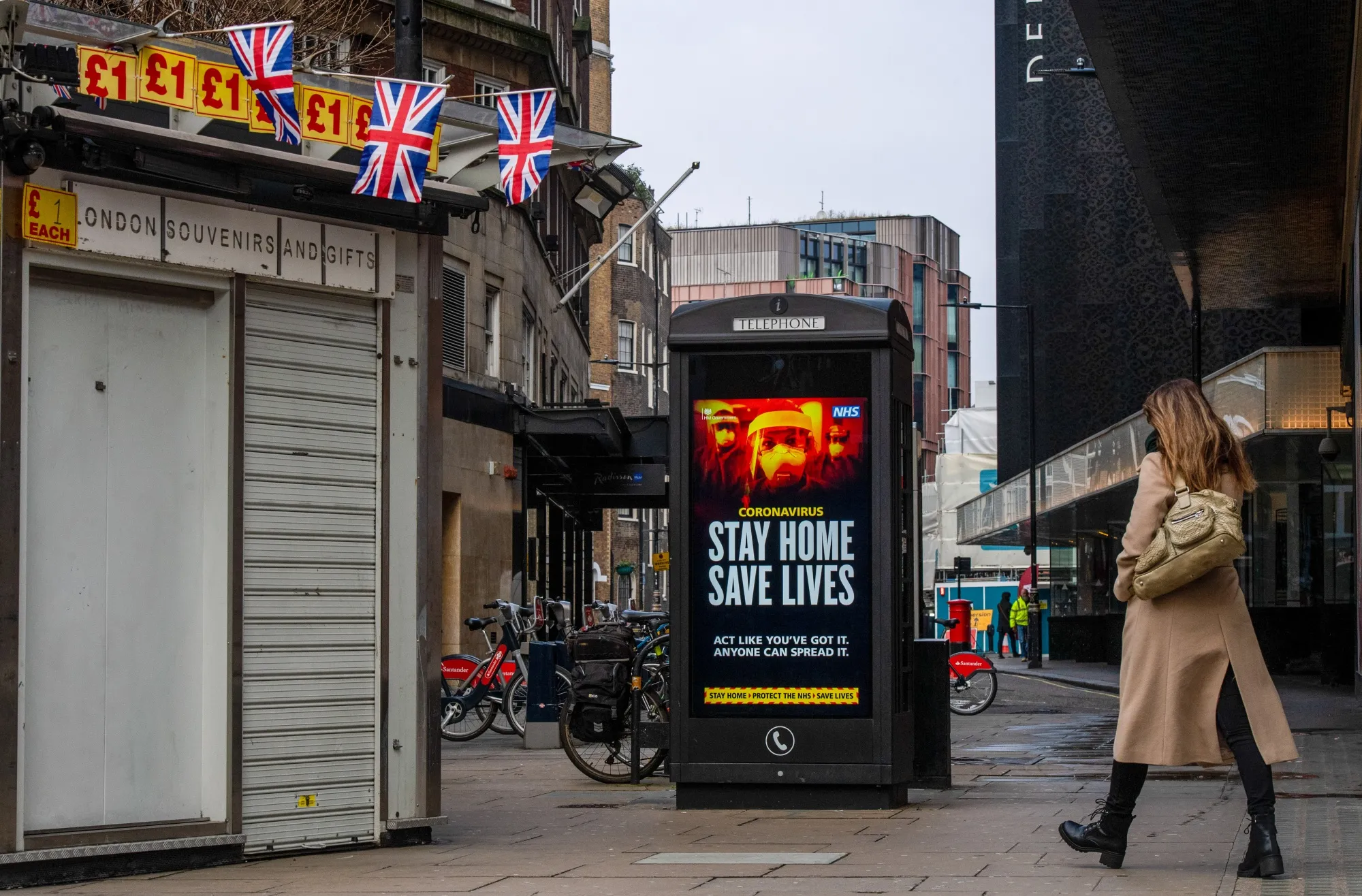 A woman walks past a closed souvenir stall and government messaging on a telephone box on Oxford Street in London.