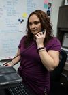 Helena Girouard sits at a desk in front of a keyboard and holds the phone receiver up to her ear. She has curly auburn hair and is wearing a plum-colored outfit. A white board and post-it notes are in the background.