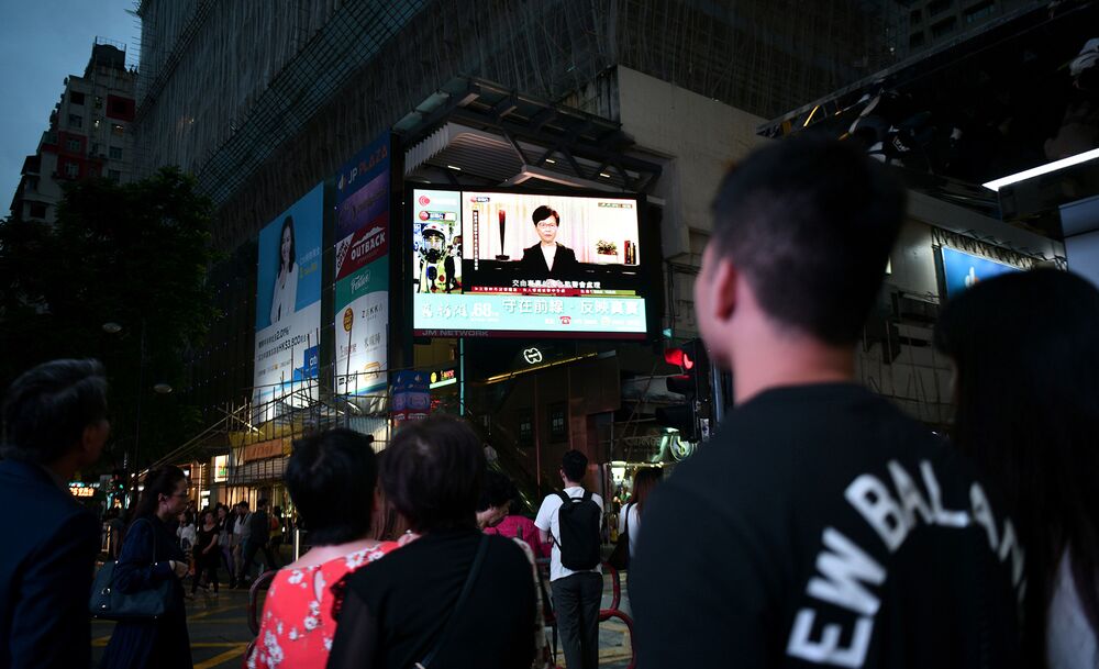 Pedestrians look at a large monitor display showing a news broadcast of Hong Kong Chief Executive Carrie Lam.