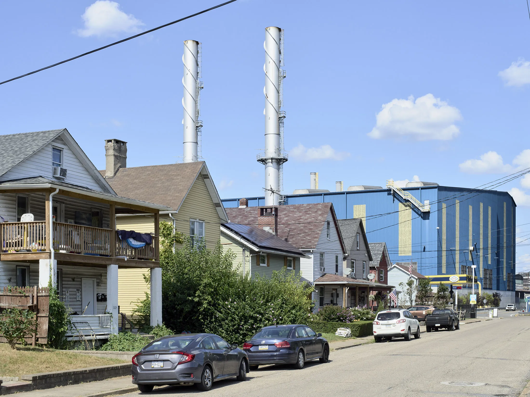 A photograph of a street with several houses in the foreground. Behind the houses is a large blue building, the Allegheny Technologies plant, with tall smokestacks extending beyond the frame of the photo.