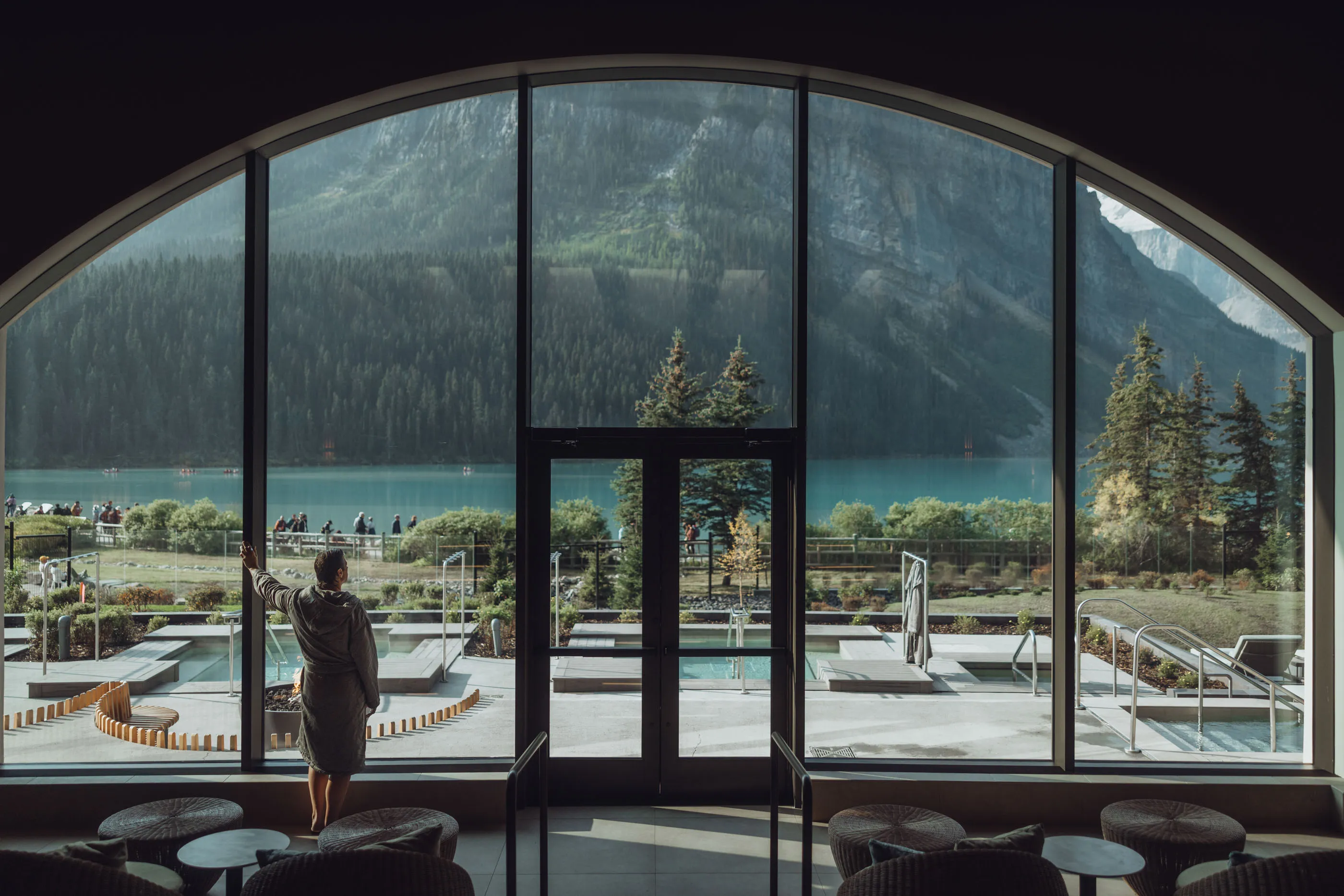 A guest stands in front of dramatic arched windows at Basin Glacial Waters, a thermal spa facility at Fairmont Chateau Lake Louise with views of the Canadian Rockies.