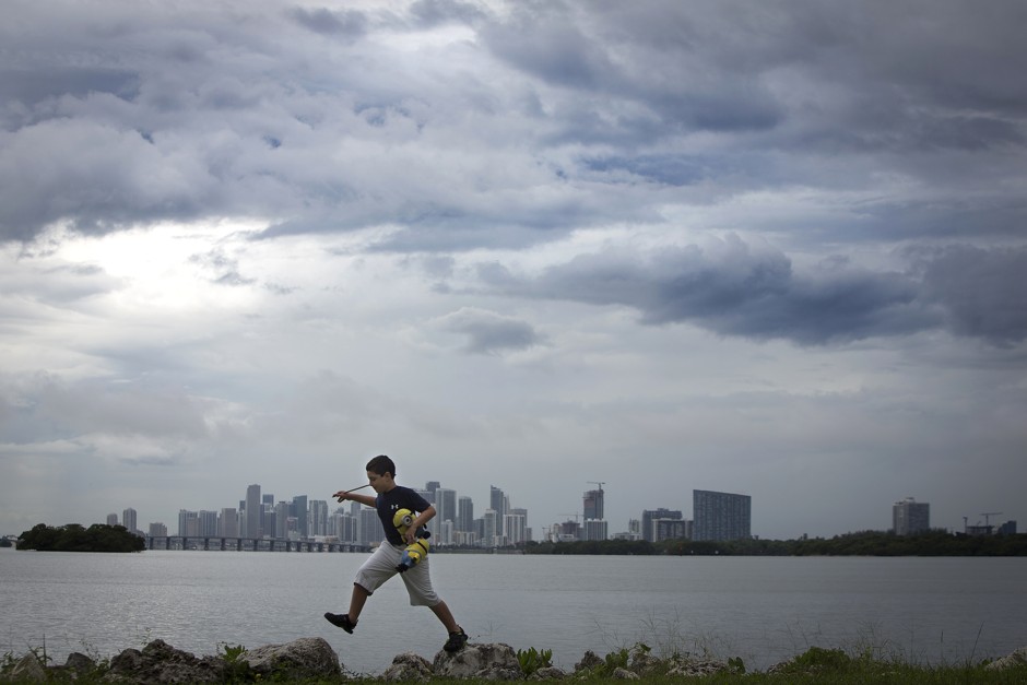 A boy jumps on rocks as he plays along a causeway on a rainy day with the city skyline in background in Miami, Florida.