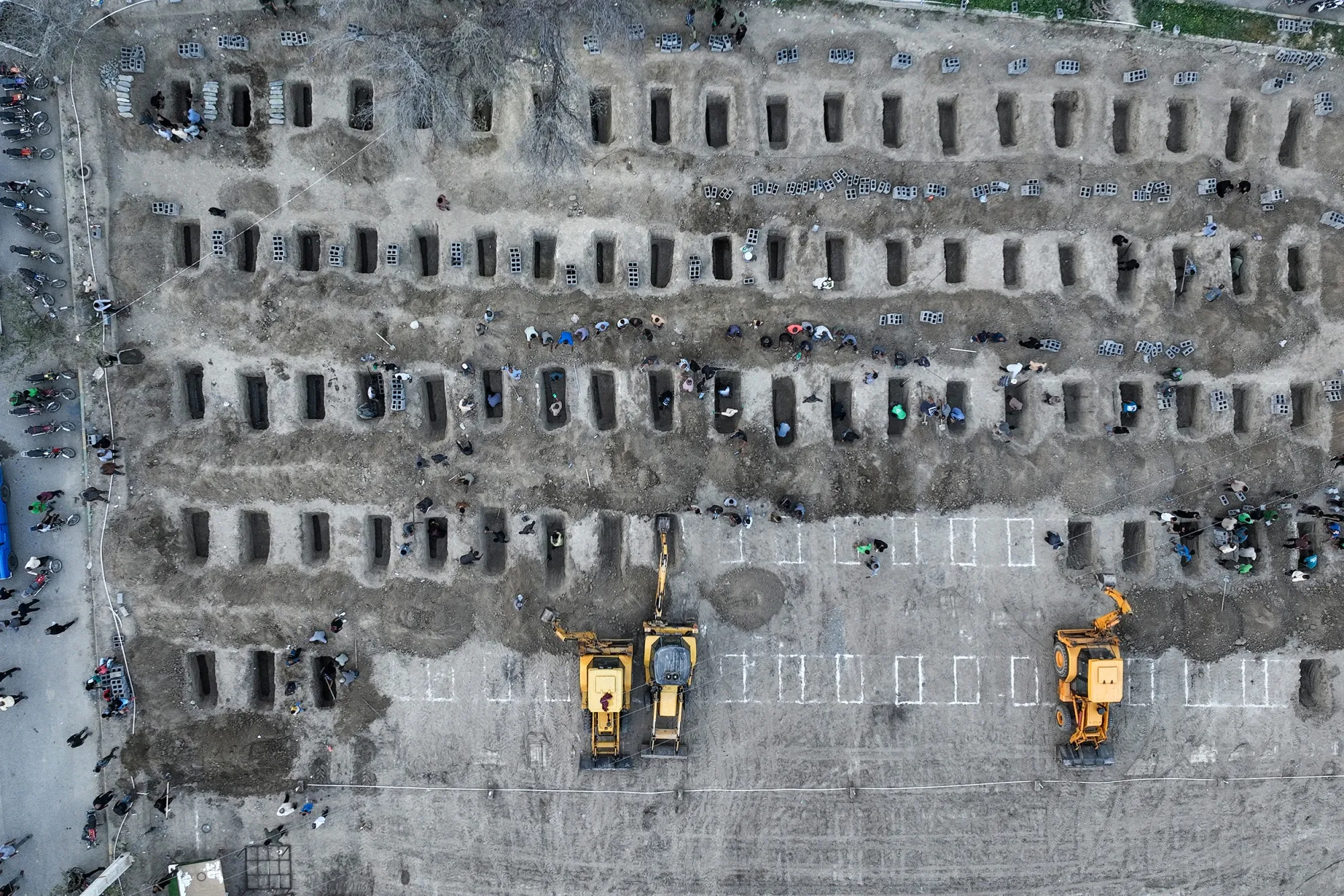 An aerial image shows rows of open graves with people by them. In one corner of the image, white lines mark undug graves, while three backhoes are working to dig.